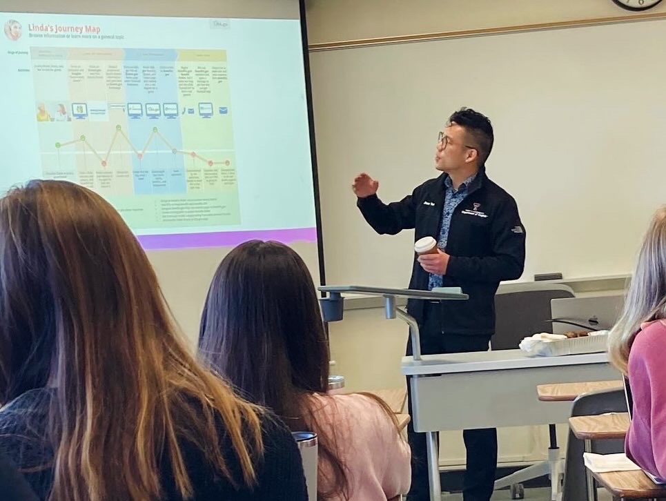 Jason gesturing and standing at the front of the classroom, next to a projector screen.